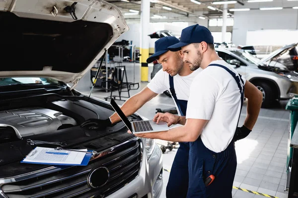 Meccanica auto guardando lo schermo del computer portatile vicino al cofano aperto dell'auto alla stazione di servizio — Foto stock