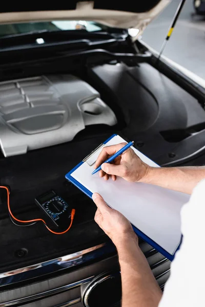 Cropped view of auto mechanic writing on clipboard near open hood of car with multimeter at service station — Stock Photo