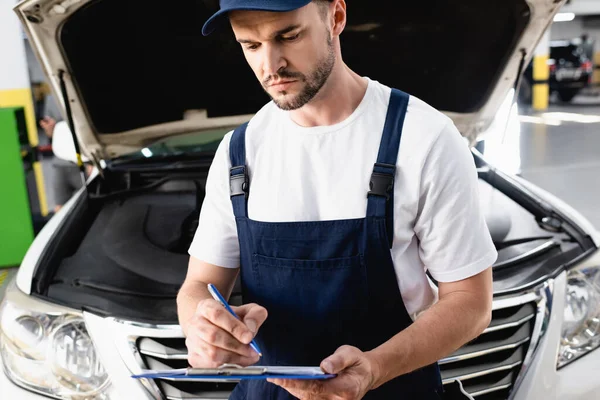 Automechaniker schreibt auf Klemmbrett nahe geöffneter Motorhaube von Auto an Tankstelle — Stockfoto