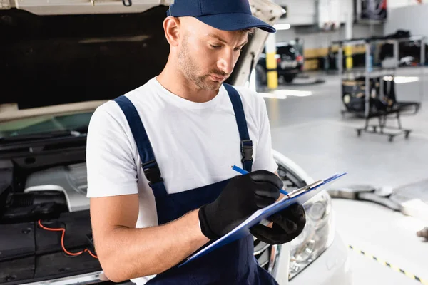 Auto mechanic writing on clipboard near open hood of car at service station — Stock Photo