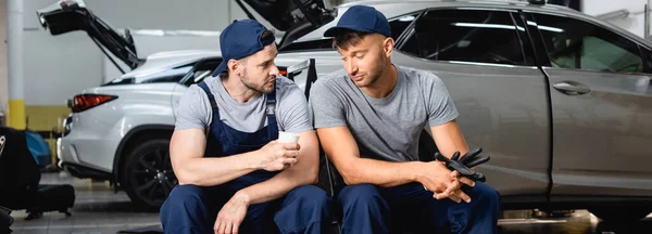 Panoramic shot of tired auto mechanic sitting near coworker and holding paper cup at service station — Stock Photo