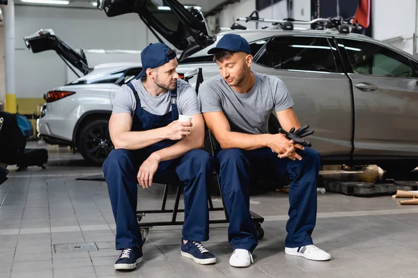 Tired auto mechanic sitting near coworker and holding paper cup at service station — Stock Photo