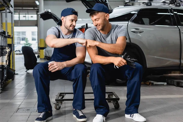 Smiling auto mechanic giving fist bump to colleague near autos at service station — Stock Photo