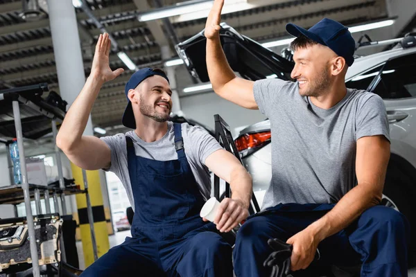 Joyeux mécanicien automobile assis et donnant cinq élevé à un collègue à la station-service — Photo de stock