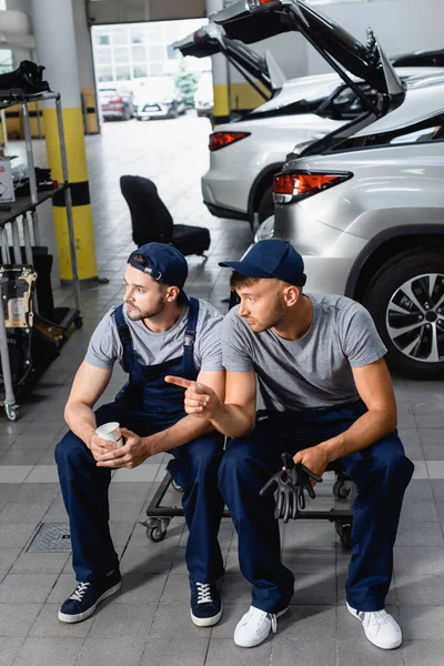 Auto mechanic sitting and holding paper cup near colleague pointing with finger at service station — Stock Photo