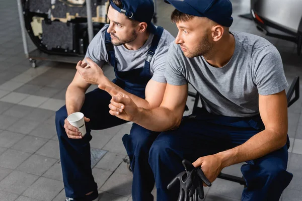 Automechaniker sitzt mit Pappbecher neben Kollege und zeigt mit Finger auf Tankstelle — Stockfoto