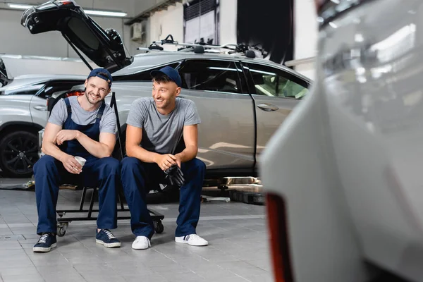 Selective focus of happy auto mechanics sitting and holding paper cup and latex gloves at service station — Stock Photo