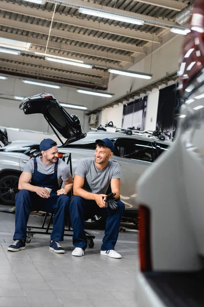 Selective focus of happy auto mechanics sitting and holding paper cup and latex gloves at service station — Stock Photo