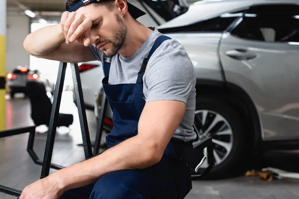 Mecánico de automóviles cansado con la mano cerca de la frente sentado cerca de los coches en la estación de servicio — Stock Photo