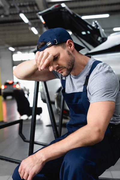 Cansado mecânico de automóveis com a mão perto da testa sentado perto de carros na estação de serviço — Fotografia de Stock