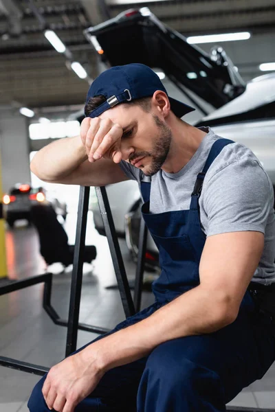 Mecánico de automóviles cansado con la mano cerca de la frente sentado cerca de los coches en la estación de servicio — Stock Photo