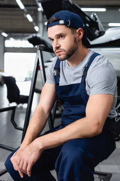 Tired auto mechanic sitting near cars at service station — Stock Photo