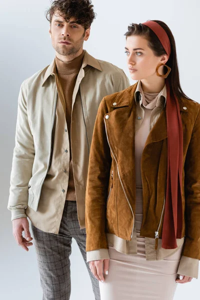 Hombre de moda mirando a la cámara cerca de chica atractiva en chaqueta de gamuza con estilo aislado en gris - foto de stock