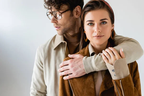 Homem elegante em óculos abraçando mulher elegante olhando para a câmera em cinza — Fotografia de Stock