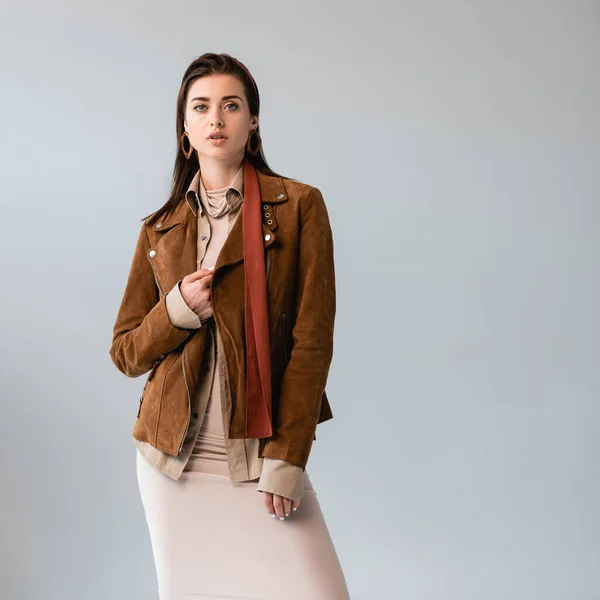 Attractive, fashionable girl touching suede jacket while looking at camera isolated on grey — Stock Photo