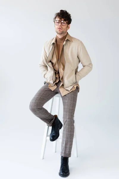 Vue pleine longueur de l'homme à la mode assis sur le tabouret, tenant les mains dans les poches et regardant la caméra sur le gris — Photo de stock