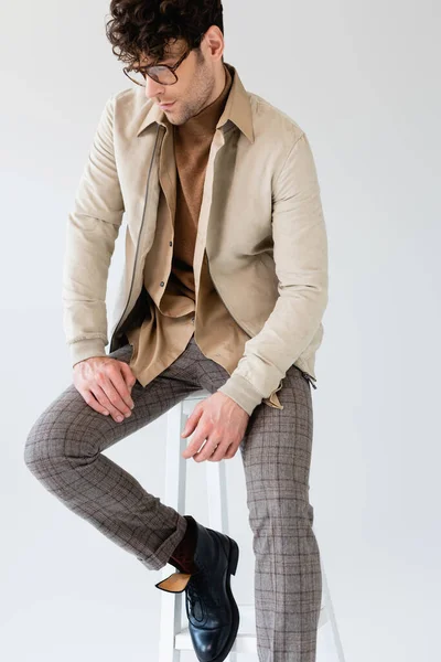 Beautiful man in stylish autumn clothes sitting on stool isolated on grey — стоковое фото