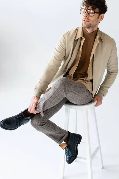 Stylish man in eyeglasses sitting on stool with crossed legs and looking away on grey — Stock Photo