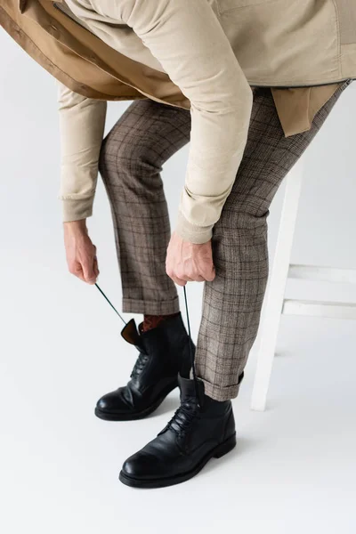 Cropped view of fashionable man tying laces on black boots while sitting on stool on white — Stock Photo