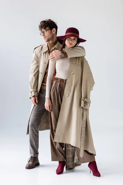Full length view of trendy man hugging stylish woman while posing in autumn clothes on grey — Stock Photo