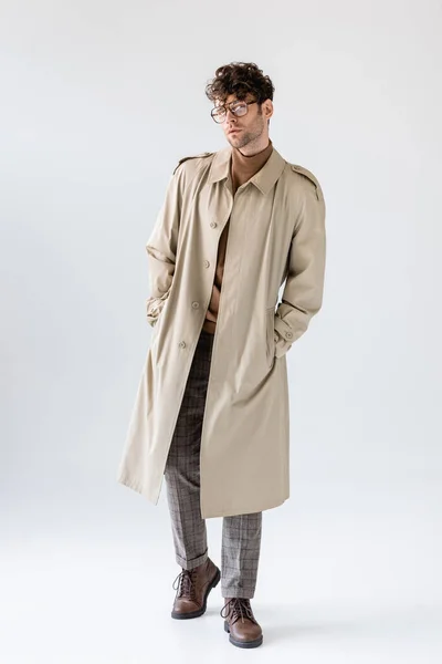 Вид в полный рост модного мужчины в трикотажном пальто, держащего руки в карманах и позирующего на сером — стоковое фото