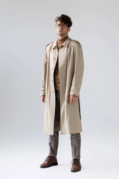 Vue pleine longueur de l'homme à la mode en trench coat regardant la caméra tout en posant sur le gris — Photo de stock