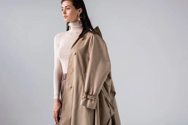Atraente, mulher na moda com casaco de trincheira no ombro posando isolado no cinza — Fotografia de Stock