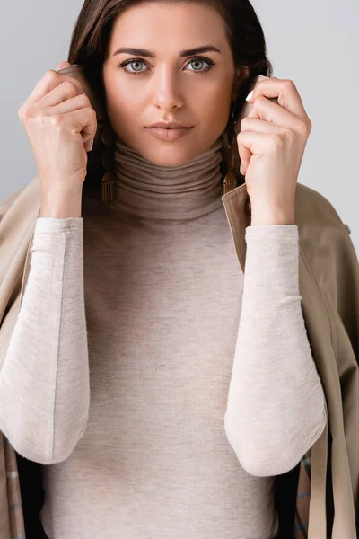 Beautiful girl touching collar of trench coat and looking at camera isolated on grey — Stock Photo