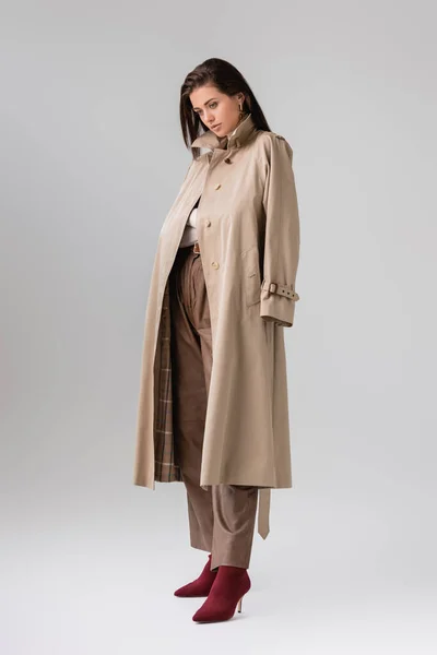 Visão de comprimento total da menina elegante com casaco de trincheira nos ombros posando em cinza — Fotografia de Stock