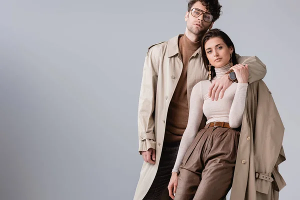 Homme à la mode en trench coat étreignant femme à la mode tout en regardant la caméra isolée sur gris — Photo de stock