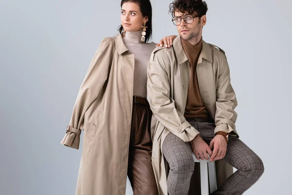 Trendy woman looking away while touching shoulder of stylish man sitting on stool isolated on grey — Stock Photo