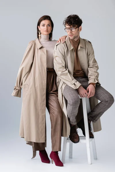 Vista completa de la mujer de moda tocando hombro de hombre de moda sentado en gris - foto de stock