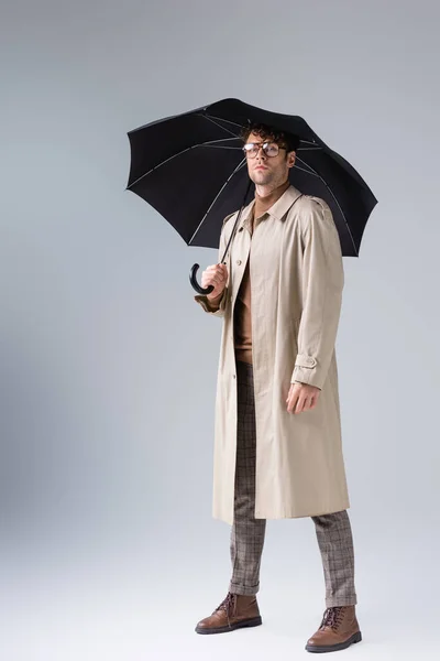 Full length view of confident, stylish man looking at camera while posing under umbrella on grey — Stock Photo