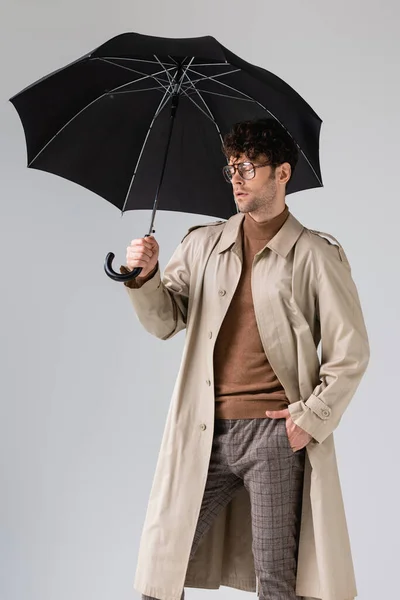 Stylish man standing under black umbrella with closed eyes isolated on grey — Stock Photo