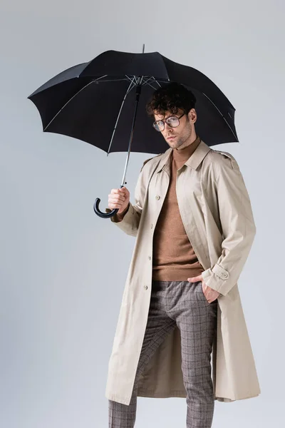 Confident, trendy man standing under umbrella with hand in pocket on grey — Stock Photo