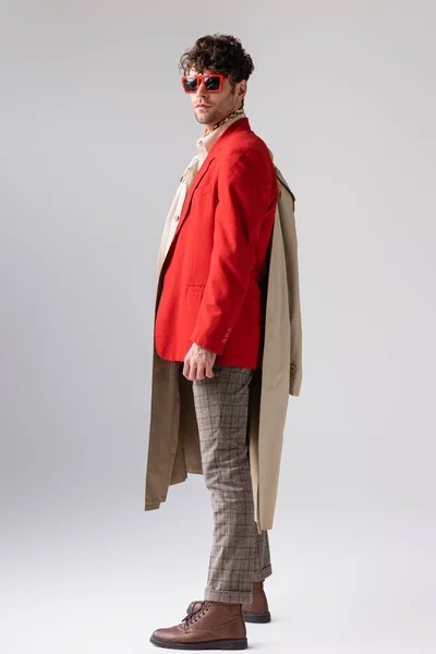 Vista completa de hombre elegante en chaqueta roja sosteniendo gabardina y mirando a la cámara en gris - foto de stock