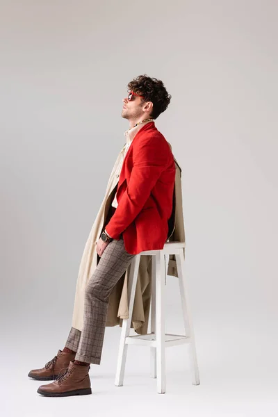 Vista lateral del joven en traje de otoño de moda mirando hacia otro lado mientras se apoya en el taburete en gris - foto de stock