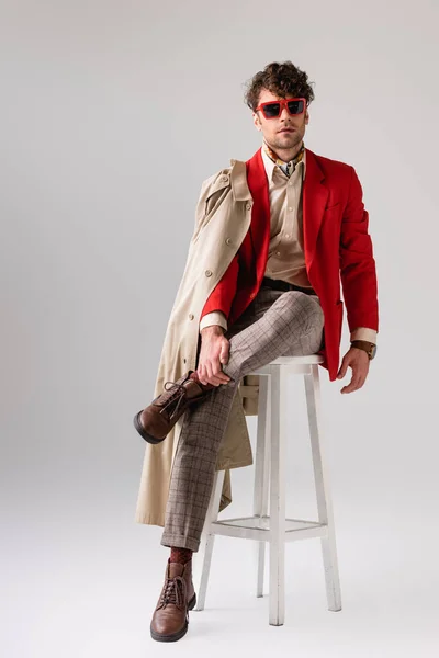 Handsome, confident man in stylish autumn clothes touching leg while sitting on stool on grey — Stock Photo