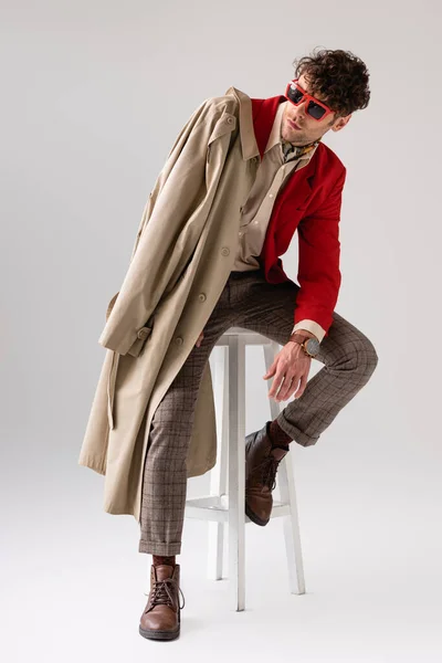 Stylish man in sunglasses sitting on stool with trench coat over shoulder and looking away on grey — Stock Photo
