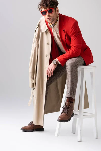 Modischer Mann im roten Blazer mit Trenchcoat auf der Schulter sitzt auf grau — Stockfoto