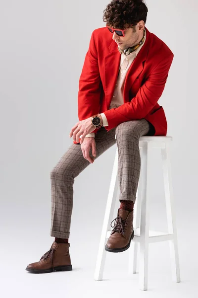 Homme à la mode en rouge blazer et lunettes de soleil assis sur tabouret avec la tête inclinée sur gris — Photo de stock