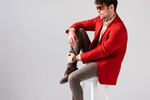 Hombre elegante en chaqueta roja y gafas de sol tocando la pierna mientras está sentado en el taburete aislado en gris - foto de stock