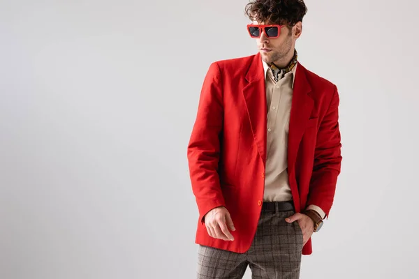 Модный мужчина в красном блейзере и солнечных очках, держась за руку в кармане, изолированном на сером — стоковое фото