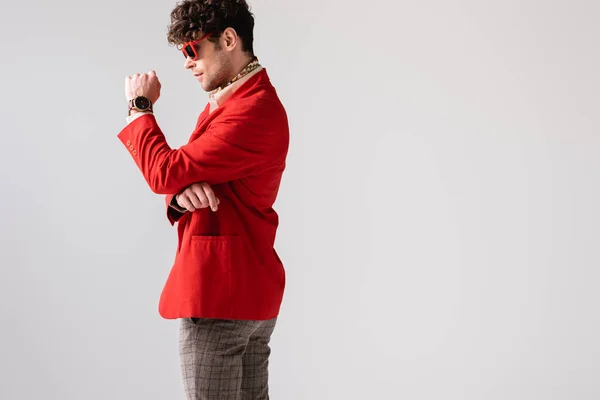 Вид сбоку стильного мужчины в красном блейзере и солнцезащитных очках, позирующего изолированным на сером — стоковое фото
