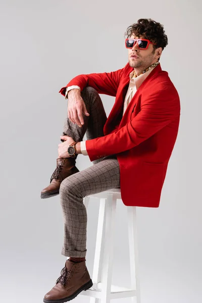 Hombre de moda en chaqueta roja y gafas de sol tocando la pierna mientras está sentado en el taburete aislado en gris - foto de stock