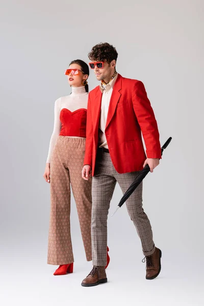 Full length view of fashionable man with folded umbrella walking near stylish woman on grey — Stock Photo