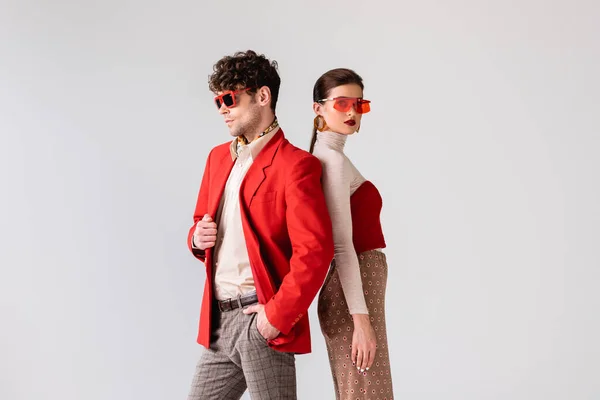 Fashionable man with hand in pocket standing back to back with stylish woman isolated on grey — Stock Photo