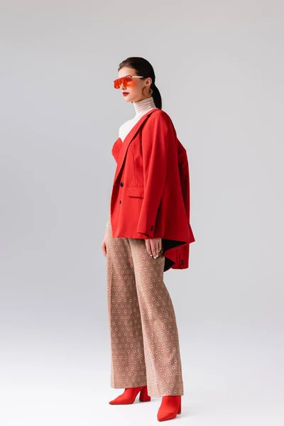 Full length view of beautiful, stylish girl with red blazer on shoulder looking away on grey — Stock Photo