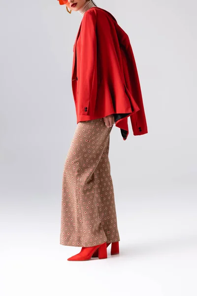 Обрізаний вид елегантної дівчини в бежевих брюках і червоний блейзер на плечах позує на сірому — стокове фото