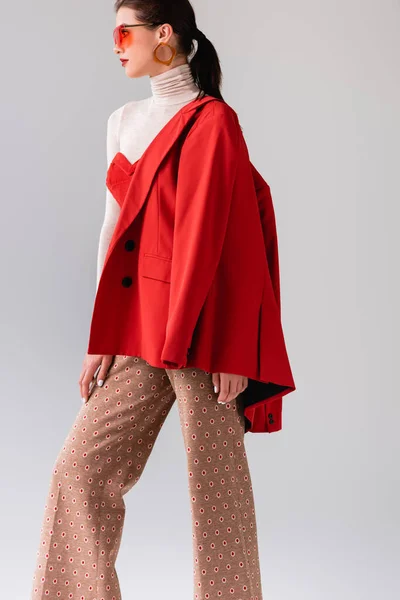 Elegante chica con chaqueta roja en el hombro mirando hacia otro lado mientras posando aislado en gris - foto de stock